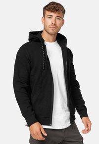 Black zip-up hoodie with a drawstring hood, front pockets, ribbed cuffs, and a textured fabric. Worn over a white T-shirt.