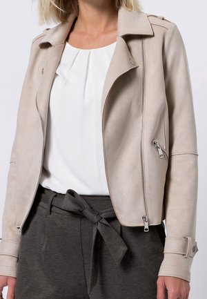 Femme portant une veste en daim beige sur un chemisier blanc et un pantalon taille haute gris foncé avec ceinture nouée, debout devant un fond uni.