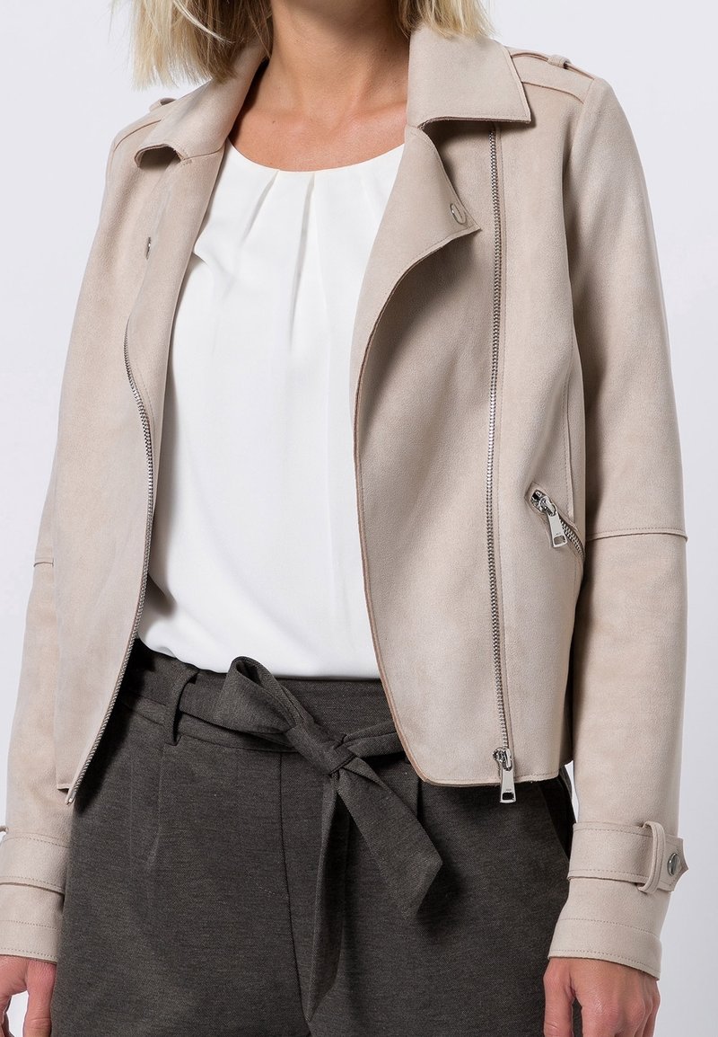 Femme portant une veste en daim beige sur un chemisier blanc et un pantalon taille haute gris foncé avec ceinture nouée, debout devant un fond uni.