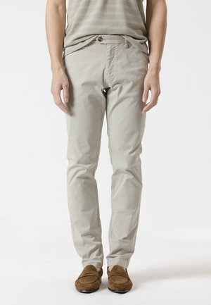 Persona vistiendo pantalones beige de corte recto, una blusa texturizada beige claro y mocasines de ante marrón, de pie frente a un fondo blanco liso.