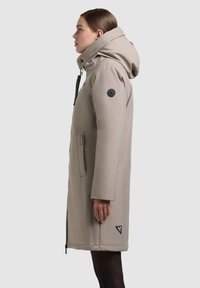 Béžový prešívaný kabát s odnímateľnou kapucňou, vybavený zipsom a bočnými vreckami. Obsahuje čierny logo nášivku na rukáve. Hladká textúra.