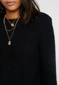 Frau mit einem schwarzen strukturierten Strickpullover und geschichteten goldenen Halsketten mit Anhänger- und Kettendetails.