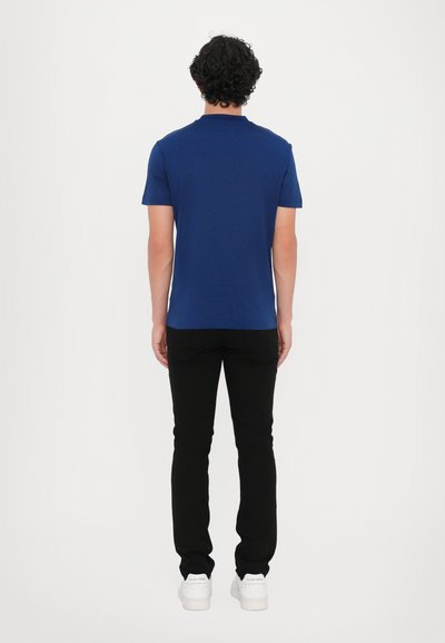 Tamsiai mėlynas trumpas rankovės marškinėlis, medvilnės mišinys, apvaliu iškirpimu, derinamas su juodomis lieknomis kelnėmis ir baltomis sportbačiais, rodomas iš nugaros.