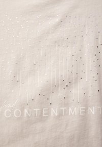 Beige t-shirt med silver- och svarta paljetter arrangerade i vertikala linjer ovanför frasen "hitta TILLFRIHET" i vit text.