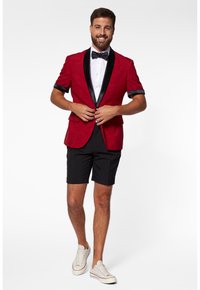 Crvena smoking jakna s crnim satenskim reverima, nošena preko bijele košulje i crnih kratkih hlača. Model nosi bijele tenisice, smiješi se, ima kratku kosu.