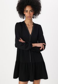 Robe noire avec de longues manches bouffantes, un décolleté en V profond et des plis verticaux. Présente des clous en argent le long de l'encolure et des boutons.