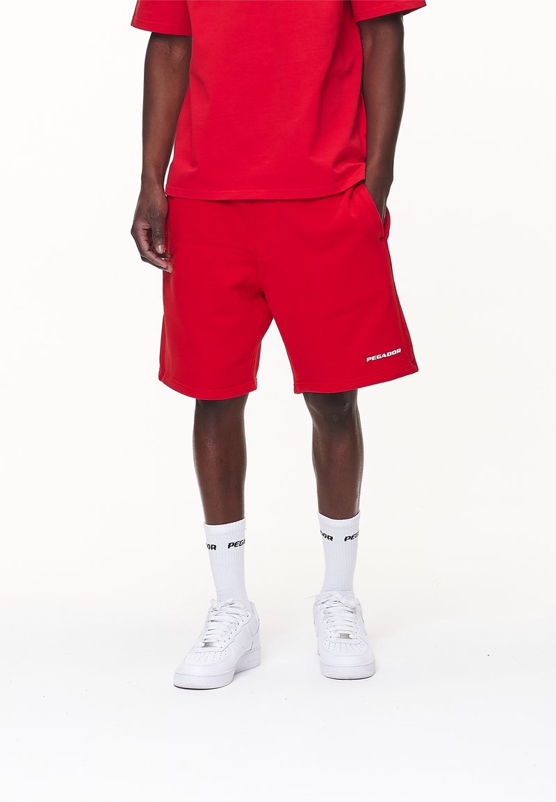 Shorts de sport rouges en tissu léger, avec poches latérales, taille élastique et logo blanc. Associés à un t-shirt rouge et des baskets blanches.