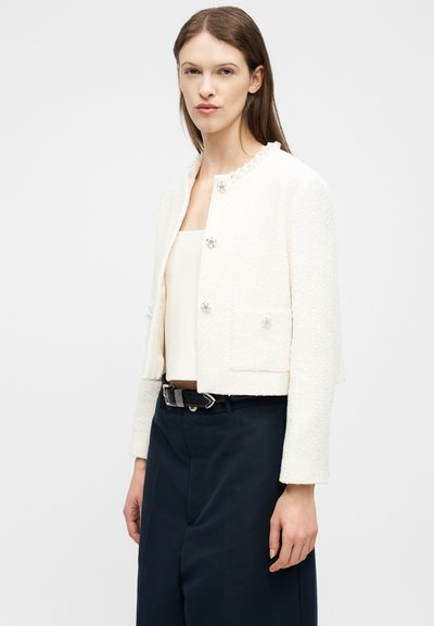 Γυναίκα που φοράει λευκό υφασμάτινο κοντό σακάκι με κουμπιά από πέρλες και λευκό τοπ, συνδυασμένο με ψηλόμεσα σκούρα παντελόνια και μαύρη ζώνη.