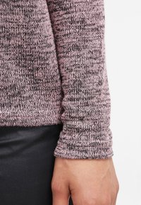 Gros plan d'une main à côté d'un poignet à manches longues en maille chinée rose et noire, porté au-dessus d'un pantalon en tissu noir.