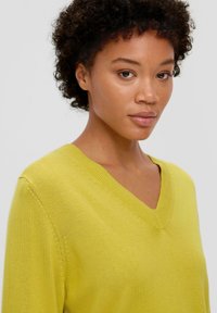 Hellgelber V-Ausschnitt-Pullover aus weichem Material mit glatter Textur. Verfügt über dezente Nahtdetails an den Schultern.