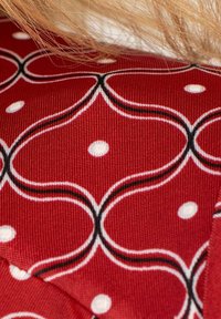 Punainen kangas, jossa on geometrinen kuvio päällekkäisistä ympyröistä, mustalla ääriviivalla ja valkoisilla pisteillä, korostaen teksturoitua kudontaa.