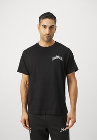 Melnas kokvilnas T-krekls ar īsām piedurknēm, kuram uz kreisā krūts ir logotips. Modelis to valkā ar tumši pelēkiem biksēm.