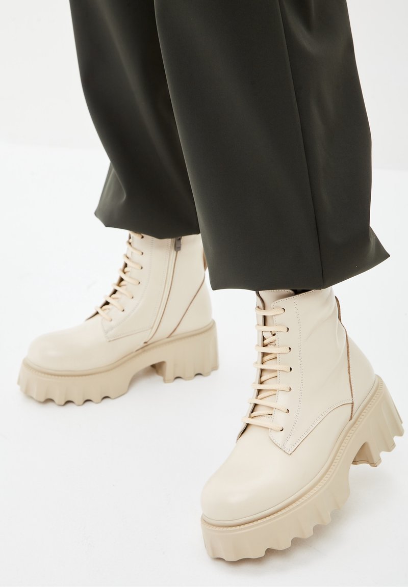 Botas de plataforma gruesas en cuero crema con frente de cordones, costuras en contraste y una suela gruesa y estriada. Combinadas con pantalones verdes oscuros.