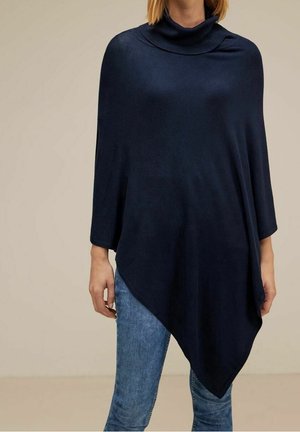 Kvinde iført en mørkeblå asymmetrisk poncho med en rullekrave, kombineret med blå jeans, stående mod en ensfarvet beige baggrund.