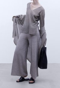 Top grigio chiaro lavorato a maglia con maniche larghe, abbinato a pantaloni coordinati a vestibilità ampia. Una borsa a tracolla nera e sandali piatti completano l'outfit.