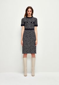 Vestido de punto en un patrón texturizado en blanco y negro, con mangas cortas, dos bolsillos en el pecho con botones dorados y una silueta ajustada.