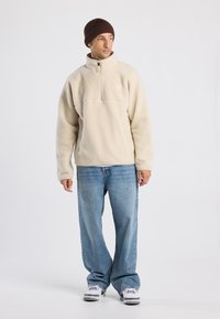 Pullover in fleece beige con zip a metà, dotato di un piccolo logo, abbinato a jeans a gamba larga di un azzurro chiaro e sneakers bianche, completato da un berretto a coste marrone.