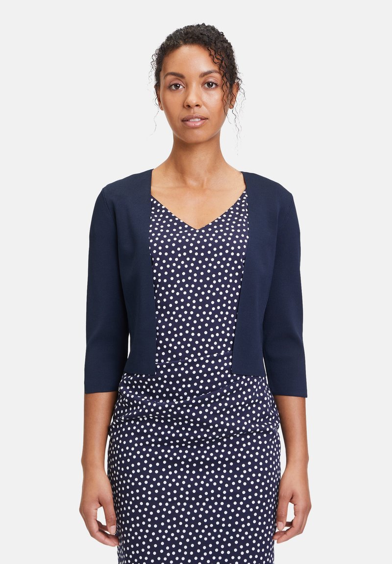 Donna con capelli ricci indossa un vestito blu navy a pois e un cardigan corto blu navy con maniche a tre quarti su uno sfondo semplice.