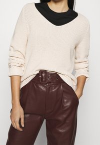 Femme portant un pull en maille crème sur un haut noir, associé à un pantalon en cuir marron foncé taille haute avec une ceinture large.