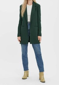 Mörkgrön kavaj med skräddarsydd passform, slagkrage och två framfickor, i kombination med ljusblå raka jeans och beiga klackstövlar.