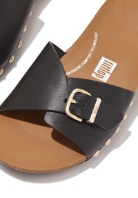Zwarte slide sandaal met een brede band en gouden gespdetails. Bruin gevoerd voetbed en tan zool met kleine ronde studs langs de rand.