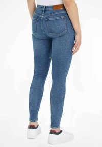 Jeans de denim azul de corte ajustado con una cintura alta, bolsillos traseros y una parche de cuero marrón. El material parece elástico y duradero.