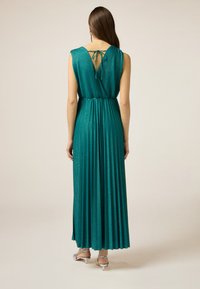 Teal-farbenes, plissiertes Maxikleid mit einer Bindeschleife und dezentem Glanz. Hat eine taillierte Taille und einen fließenden Rock für Bewegungsfreiheit. Silberne High Heels Sandalen.