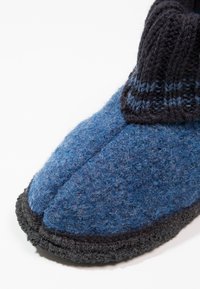 Pantoufle en feutre de laine avec un dessus bleu, un poignet côtelé sombre et une semelle en caoutchouc noir texturé. La vue rapprochée montre les détails de la couture et du matériau.