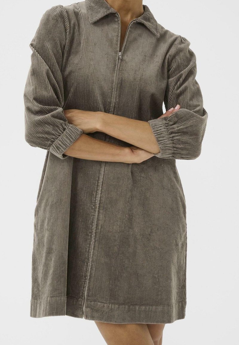 Femme portant une robe en velours côtelé taupe avec manches trois-quarts et fermeture éclair devant, bras croisés sur la poitrine, debout devant un fond blanc.