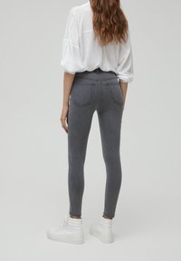 Femme aux cheveux longs et ondulés, portant un jean skinny gris, un chemisier blanc à manches retroussées et des baskets blanches montantes, debout et tournée de dos.