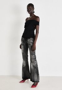 Svart avskjært topp, slitne grå detaljerte flared jeans, og glitrende røde stiletthæler. Modellen står med armene avslappet ved siden av seg.