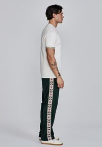 Camisa blanca de manga corta con un puño de brazo con logo, combinada con pantalones de chándal verdes que tienen una franja floral en el costado y zapatillas blancas.