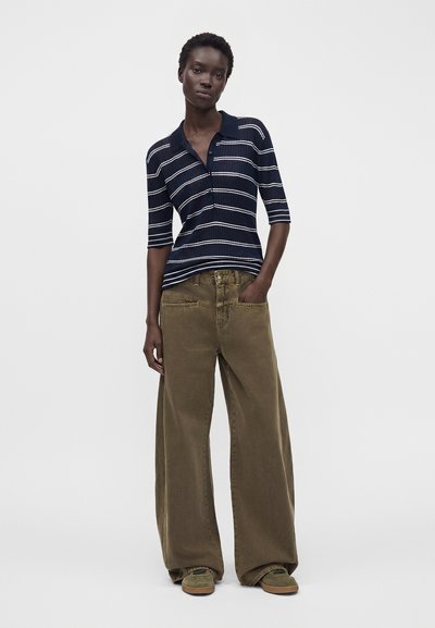 Femme debout portant un polo bleu marine rayé à manches courtes, un pantalon marron à jambes larges et des chaussures vertes, avec une main dans la poche.