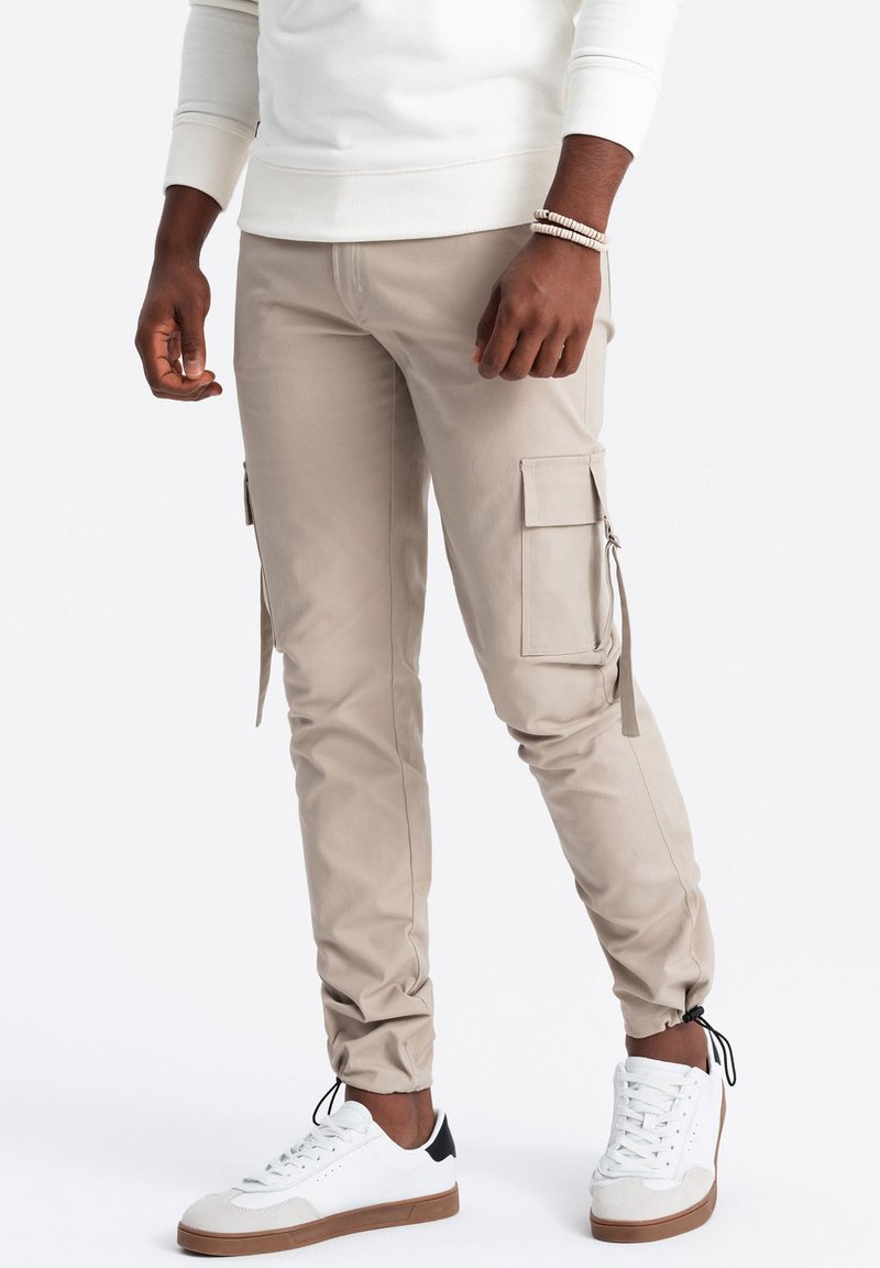 Béžové cargo kalhoty slim fit s několika kapsami, zipy a elastickými manžetami. V kombinaci s bílými teniskami a světlým topem.