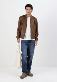 Brun bomberjakke med quiltet tekstur, hvid t-shirt, blå slidte jeans, beige fodtøj og en totebag.