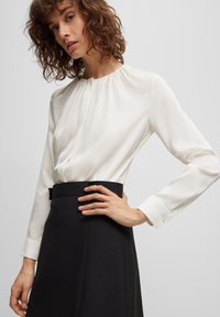 Weiße Bluse mit langen Ärmeln und plissiertem Design, kombiniert mit einem schwarzen, hoch sitzenden Rock. Glattes Gewebe und figurbetonte Silhouette.