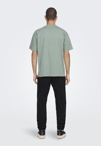 Groene katoenen T-shirt met korte mouwen, ontspannen pasvorm, gecombineerd met zwarte broek en sneakers met witte zolen en paarse accenten.