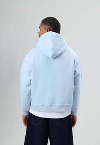 Sweat à capuche court bleu clair avec capuche à cordon, poignets et ourlet côtelés. Texture lisse, porté sur une chemise blanche et associé à un jean foncé.