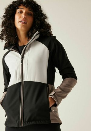 Frau mit lockigem Haar, die eine schwarz-weiße und beige Reißverschlussjacke trägt, mit den Händen in den Taschen vor einem schlichten Hintergrund.