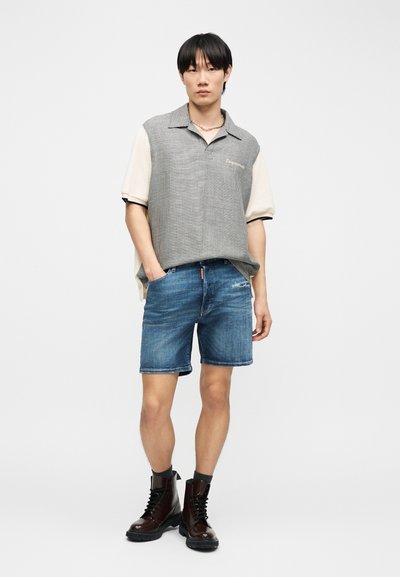 Jeune homme debout les mains dans les poches, portant une chemise à manches courtes, un short en jean et des bottes sombres à lacets sur un fond uni.