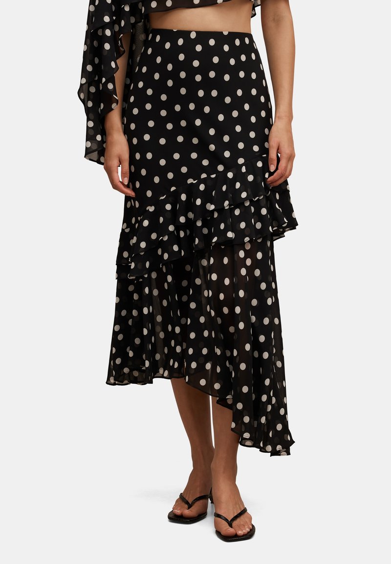 Fustă midi asimetrică neagră cu buline albe și tiv stratificat cu volane, purtată cu sandale negre cu toc și barete.