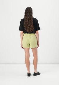 Sort t-shirt, korte limegrønne shorts med elastisk talje og sorte flade sko. Lang bølget hår set fra bagenden.