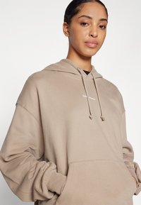 Beige hoodie met een losse pasvorm, een trekkoordcapuchon en een voorkantzak. Het materiaal lijkt zacht met zichtbare stiksels en een klein logo.