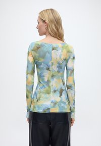 Γυναίκα με ξανθά μαλλιά που φοράει μακρυμάνικη, εφαρμοστή φλοράλ μπλούζα σε αποχρώσεις του μπλε, πράσινου και κίτρινου, συνδυασμένη με μαύρο παντελόνι, εμφανιζόμενη από την πίσω πλευρά.