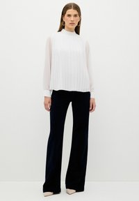 Weiße plissierte Bluse mit hohem Kragen, kombiniert mit marineblauen, ausgestellten Hosen. Die Bluse hat transparente lange Ärmel und kontrastierende Manschetten.