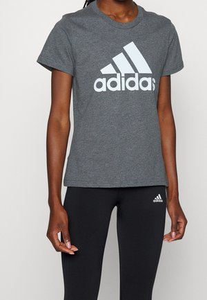 Person wearing a grey Adidas t-shirt with large logo and black Adidas leggings with small logo, standing against a plain background.