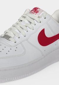 Κοντινή λήψη λευκού παπουτσιού Nike Air Force 1 με κόκκινο λογότυπο Swoosh, λευκά κορδόνια, διάτρητη μύτη και κόκκινο λογότυπο στη γλώσσα.