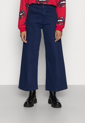 Osoba nosząca granatowe, szerokie spodnie z wysokim stanem i surowymi brzegami, czarne masywne botki i czerwoną bluzę z nadrukiem ust.