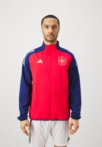 Chaqueta deportiva roja y marino con alto cuello, cremallera completa y dos bolsillos laterales. Presenta el logo de Adidas y el emblema nacional de España en el pecho.