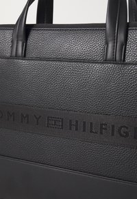 Black textured leather tote bag with flat handles. Features a smooth bottom panel and a prominent logo band across the front.